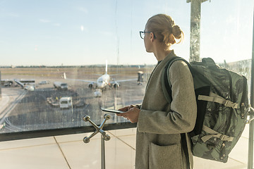 Image showing Casually dressed female traveler at airport, holding smart phone device, looking through the airport gate windows at planes on airport runway