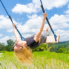 Image showing Happy female traveller swinging on wooden swing, enjoying summer vacation among pristine green rice terraces.