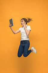 Image showing Image of young woman over orange background using laptop computer or tablet gadget while jumping.