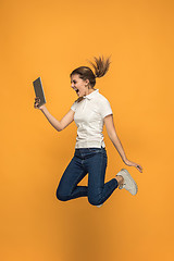 Image showing Image of young woman over orange background using laptop computer or tablet gadget while jumping.