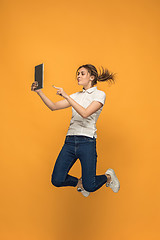 Image showing Image of young woman over orange background using laptop computer or tablet gadget while jumping.