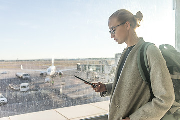 Image showing Casually dressed female traveler at airport looking at smart phone device in front of airport gate windows overlooking planes on airport runway