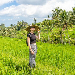 Image showing Relaxed fashionable caucasian female tourist wearing small backpack and traditional asian paddy hat walking among beautiful green rice fields and terraces on Bali island