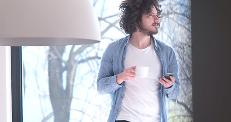 Image showing young man drinking coffee and using a mobile phone  at home