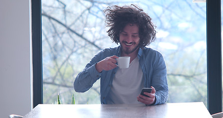 Image showing young man drinking coffee and using a mobile phone  at home