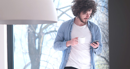 Image showing young man drinking coffee and using a mobile phone  at home