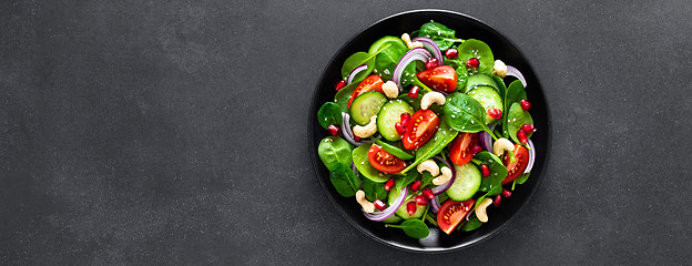 Image showing Spinach salad with fresh cucumbers, tomato, onion, pomegranate, sesame seeds and cashew nuts on black background. Healthy vegan food. Top view. Banner