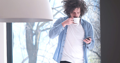 Image showing young man drinking coffee and using a mobile phone  at home