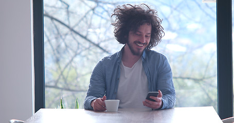 Image showing young man drinking coffee and using a mobile phone  at home