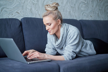 Image showing Happy blond woman lying prone on sofa and working on laptop computer