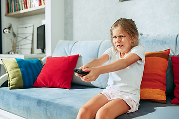 Image showing Little casual girl watching tv at home. Female kid sitting on sofa with TV remote and switching channels