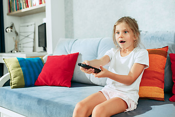 Image showing Little casual girl watching tv at home. Female kid sitting on sofa with TV remote and switching channels