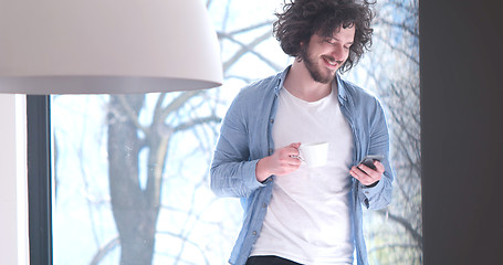 Image showing young man drinking coffee and using a mobile phone  at home