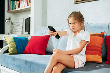 Image showing Little casual girl watching tv at home. Female kid sitting on sofa with TV remote and switching channels