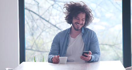 Image showing young man drinking coffee and using a mobile phone  at home