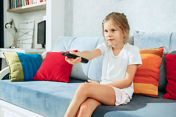 Image showing Little casual girl watching tv at home. Female kid sitting on sofa with TV remote and switching channels