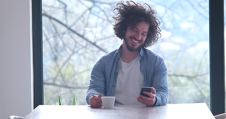 Image showing young man drinking coffee and using a mobile phone  at home