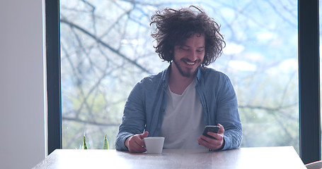 Image showing young man drinking coffee and using a mobile phone  at home