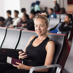 Image showing Casual tanned blond female traveler holding cell phone, passport and boarding pass while waiting to board a plane at the departure gates at the asian airport terminal