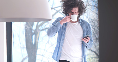 Image showing young man drinking coffee and using a mobile phone  at home