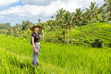 Image showing Relaxed fashionable caucasian female tourist wearing small backpack and traditional asian paddy hat walking among beautiful green rice fields and terraces on Bali island