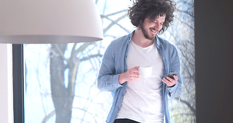 Image showing young man drinking coffee and using a mobile phone  at home