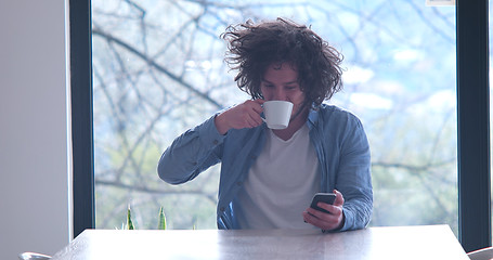 Image showing young man drinking coffee and using a mobile phone  at home