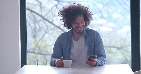 Image showing young man drinking coffee and using a mobile phone  at home