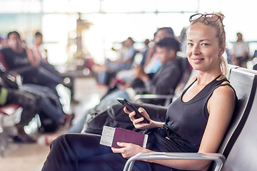 Image showing Casual tanned blond female traveler holding cell phone, passport and boarding pass while waiting to board a plane at the departure gates at the asian airport terminal
