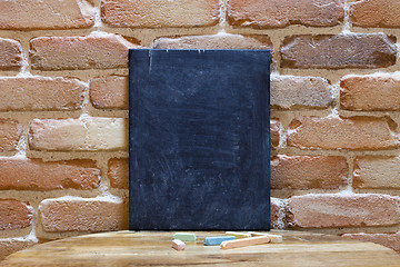 Image showing Blank blackboard on wooden table at brick wall.