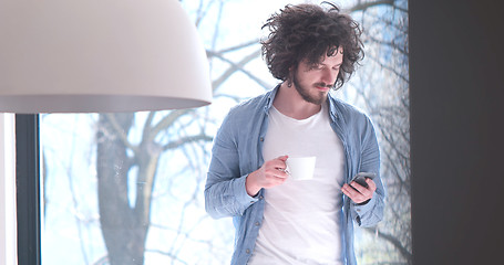 Image showing young man drinking coffee and using a mobile phone  at home