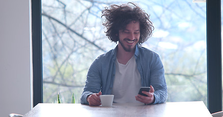 Image showing young man drinking coffee and using a mobile phone  at home