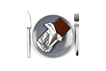 Image showing Chocolate bar with foil on blue plate, fork and knife on white b