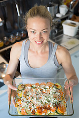 Image showing Smiling young healthy woman holding and proudly showing glass baking try with row vegetarian dish ingredients before putting it into oven.