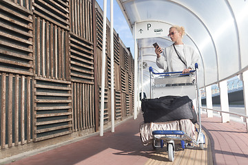 Image showing Young casual woman using mobile phone application while transporting luggage from arrival parking to international airport departure termainal by luggage trolley