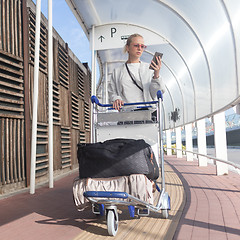 Image showing Young casual woman using mobile phone application while transporting luggage from arrival parking to international airport departure termainal by luggage trolley