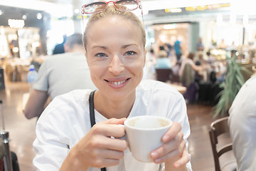 Image showing Portrait of a casual young blond woman having a cup of coffee, sitting in cafe indoors of an airport, station, food market or shopping mall.