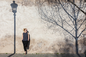 Image showing Graphical and textured artisic image of modern trendy fashionable woman wearing sunglasses leaning against old textured retro wall with vintage street lamp and tree shadow falling on the wall.