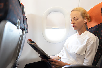 Image showing Woman reading in flight magazine on airplane. Female traveler reading seated in passanger cabin. Sun shining trough airplane window.