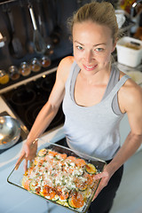 Image showing Smiling young healthy woman holding and proudly showing glass baking try with row vegetarian dish ingredients before putting it into oven.