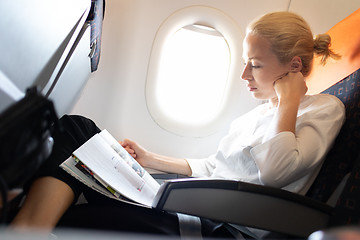 Image showing Woman reading in flight magazine on airplane. Female traveler reading seated in passanger cabin. Sun shining trough airplane window