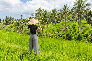 Image showing Relaxed fashionable caucasian female tourist wearing small backpack and traditional asian paddy hat looking at beautiful green rice fields and terraces on Bali island