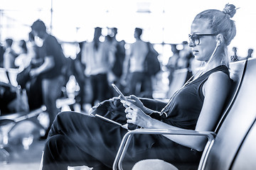 Image showing Female traveler using her cell phone while waiting to board a plane at departure gates at asian airport terminal. Blue toned black and white photo.