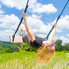 Image showing Happy female traveller swinging on wooden swing, enjoying summer vacation among pristine green rice terraces.