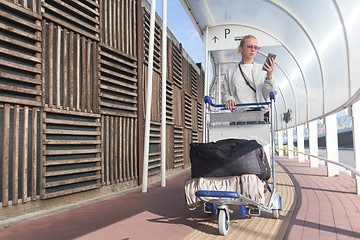 Image showing Young casual woman using mobile phone application while transporting luggage from arrival parking to international airport departure termainal by luggage trolley