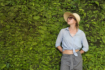 Image showing Portrait of a beautiful female traveler. Smiling young woman in summer hat wearing sunglasses, standing in front of lush tropical plant greenery wall background.