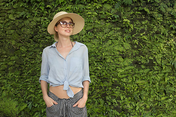 Image showing Portrait of a beautiful female traveler. Smiling young woman in summer hat wearing sunglasses, standing in front of lush tropical plant greenery wall background.