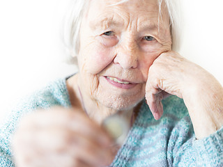 Image showing Sad elderly woman sitting at the table at home and looking miserably at only remaining coin from pension in her hand.