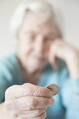 Image showing Sad elderly woman sitting at the table at home and looking miserably at only remaining coin from pension in her hand.