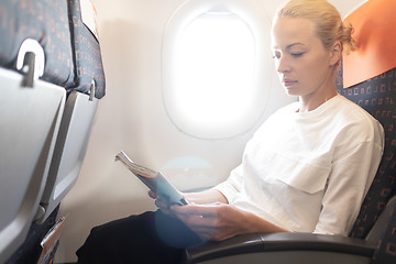 Image showing Woman reading in flight magazine on airplane. Female traveler reading seated in passanger cabin. Sun shining trough airplane window